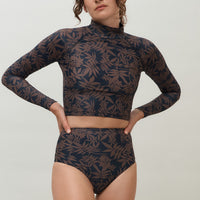 A woman wearing a high-waist sustainable swimwear bottom in a black and brown leaf print with matching long sleeve rashguard.