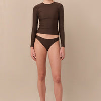 A woman wearing a brown sustainable swimwear classic brief style bikini bottom with a long sleeve rashguard.