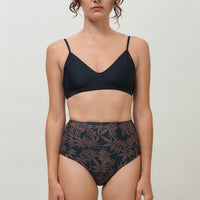 A woman wearing a high-waist sustainable swimwear bottom in a black and brown leaf print.