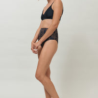 A woman wearing a high-waist sustainable swimwear bottom in a black and brown leaf print.