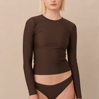 A woman wearing a brown sustainable swimwear classic brief style bikini bottom with a long sleeve rashguard.